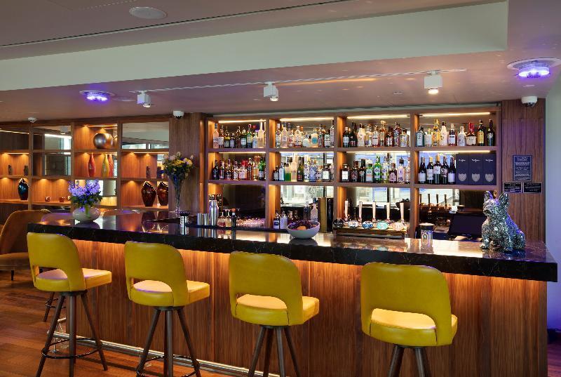 Tower Suites By Blue Orchid لندن المظهر الخارجي الصورة The photo shows a modern bar area. In the foreground, there are several yellow bar stools lined up in front of a sleek black countertop. Behind the bar, there is a well-stocked shelf displaying a variety of bottles, including spirits and liqueurs, il