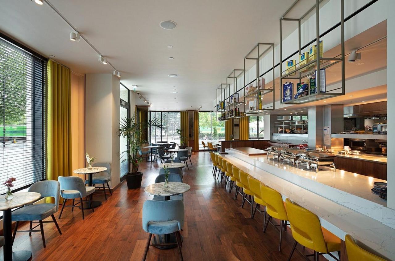 Tower Suites By Blue Orchid لندن المظهر الخارجي الصورة The photo shows a modern restaurant interior. It features a spacious layout with large windows allowing natural light to flood the space. The decor includes a combination of wooden flooring and colorful furniture—blue and yellow chairs arranged aroun