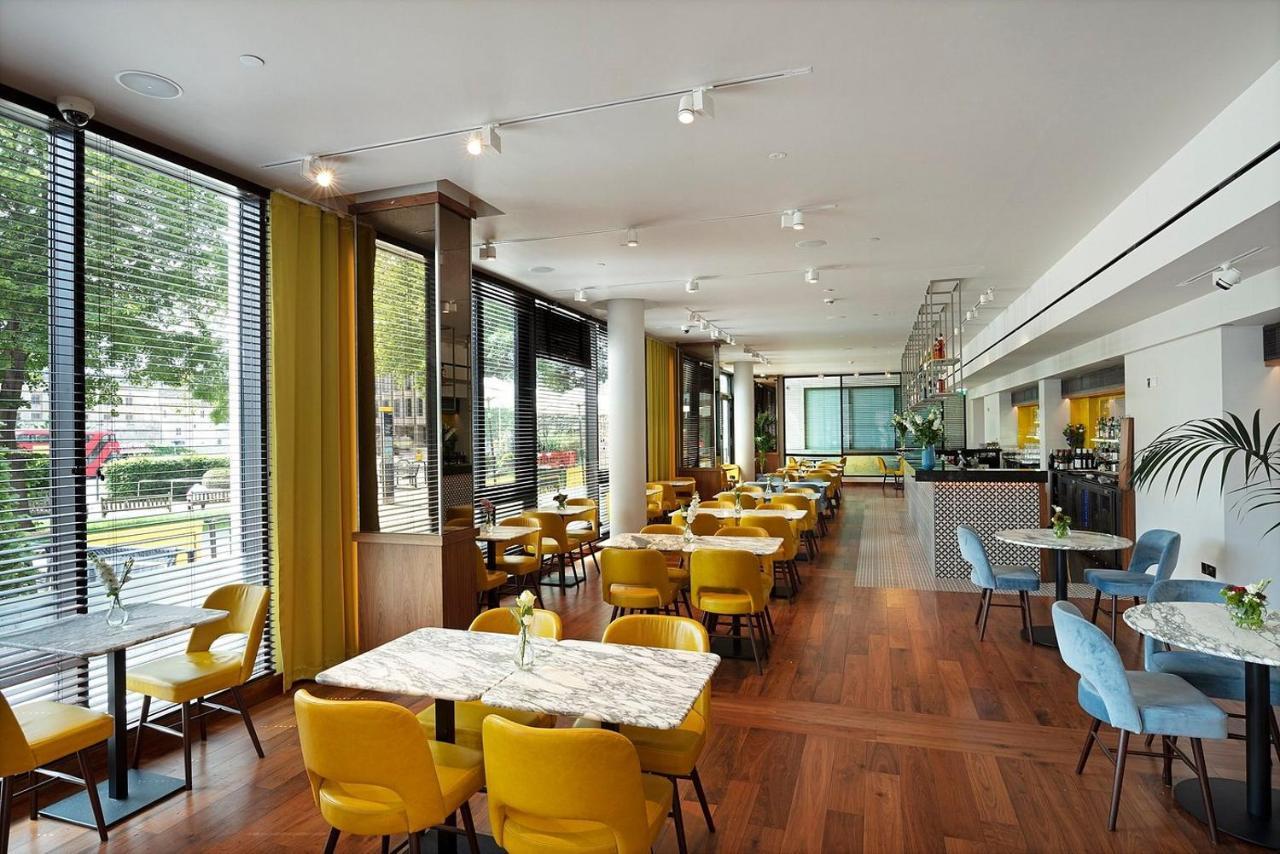 Tower Suites By Blue Orchid لندن المظهر الخارجي الصورة The photo depicts a modern dining area with a bright and airy atmosphere. There are several tables with marble tops, accompanied by yellow chairs, creating a vibrant contrast with the wood flooring. The space features large windows that allow ample n