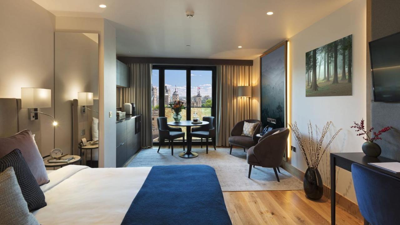 Tower Suites By Blue Orchid لندن المظهر الخارجي الصورة The photo shows a modern hotel room designed with a contemporary aesthetic. On one side, there is a large bed with a blue blanket, while the other side features a sitting area with two chairs and a small round dining table. Large windows let in natur