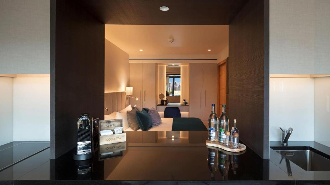 Tower Suites By Blue Orchid لندن المظهر الخارجي الصورة The image shows a modern hotel room with a stylish design. In the foreground, there is a dark countertop with several bottles displayed, likely including spirits and water. Just beyond the countertop, you can see the bedroom area, which has light-col