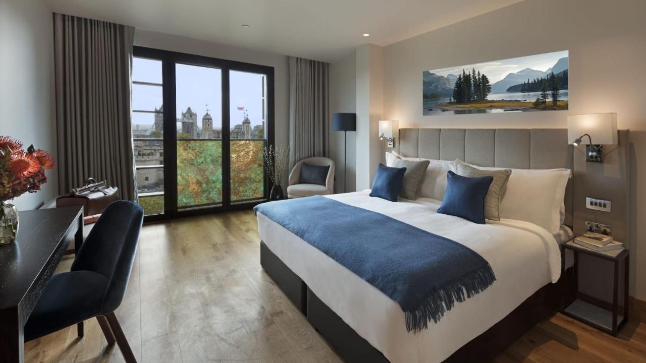Tower Suites By Blue Orchid لندن المظهر الخارجي الصورة The photo shows a modern hotel room that features a large bed with white linens and blue accent pillows. There’s a cozy throw blanket at the foot of the bed. The room is well-lit and includes a seating area with a chair and a small table. A large win
