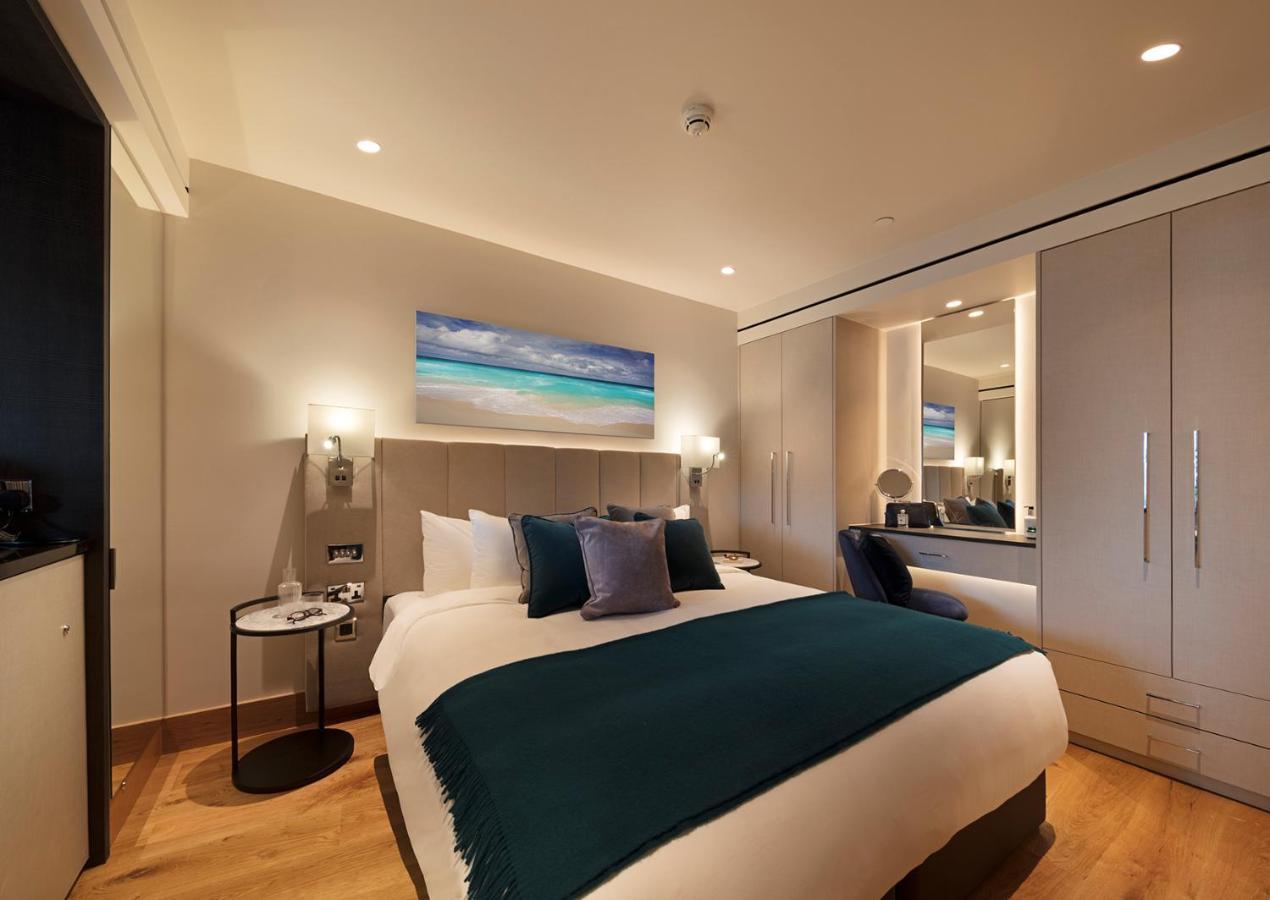 Tower Suites By Blue Orchid لندن المظهر الخارجي الصورة The photo shows a modern hotel room featuring a double bed with a white duvet and a dark teal throw blanket. There are decorative pillows in shades of blue and purple on the bed. To one side, there's a round bedside table with a small lamp. A large m