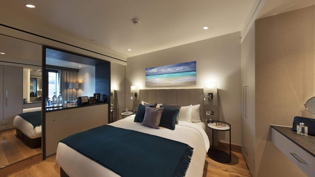 Tower Suites By Blue Orchid لندن المظهر الخارجي الصورة The photo shows a modern hotel bedroom featuring a large bed with a light-colored headboard and a deep blue throw at the foot. The bed is adorned with multiple decorative pillows. On the wall above the bed, there is a colorful painting depicting a be