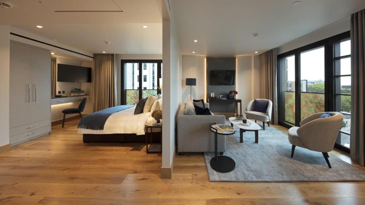 Tower Suites By Blue Orchid لندن المظهر الخارجي الصورة The photo shows a modern hotel room or suite. On one side, there's a bed with a blue and white duvet and pillows, accompanied by a bedside table with a lamp. Opposite the bed, there's a living area featuring a cozy gray sofa, a small round coffee tab