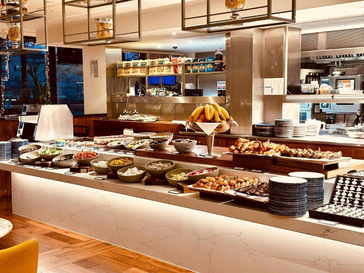 Tower Suites By Blue Orchid لندن المظهر الخارجي الصورة The photo depicts a buffet-style setup, likely in a restaurant or a hotel dining area. In the foreground, there are multiple bowls and platters filled with a variety of food items, including salads, fruits, pastries, and other dishes. There is a long