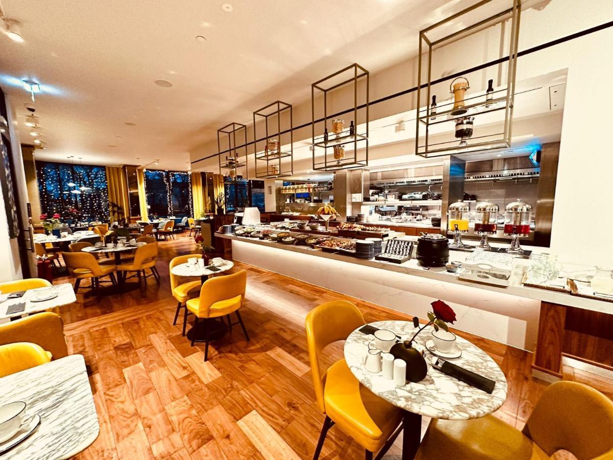 Tower Suites By Blue Orchid لندن المظهر الخارجي الصورة The photo depicts a modern restaurant interior with a spacious layout. There are several round tables, each adorned with marble tabletops and stylish yellow chairs. A buffet area is visible in the background, featuring various food setups and kitchen