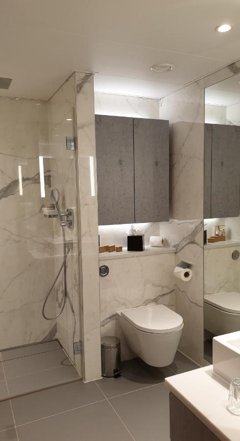Tower Suites By Blue Orchid لندن المظهر الخارجي الصورة The photo shows a modern bathroom design featuring elegant marble walls. On one side, there is a transparent shower enclosure with a showerhead and controls visible. Opposite the shower, there is a toilet, and above it are some sleek cabinets. The sp