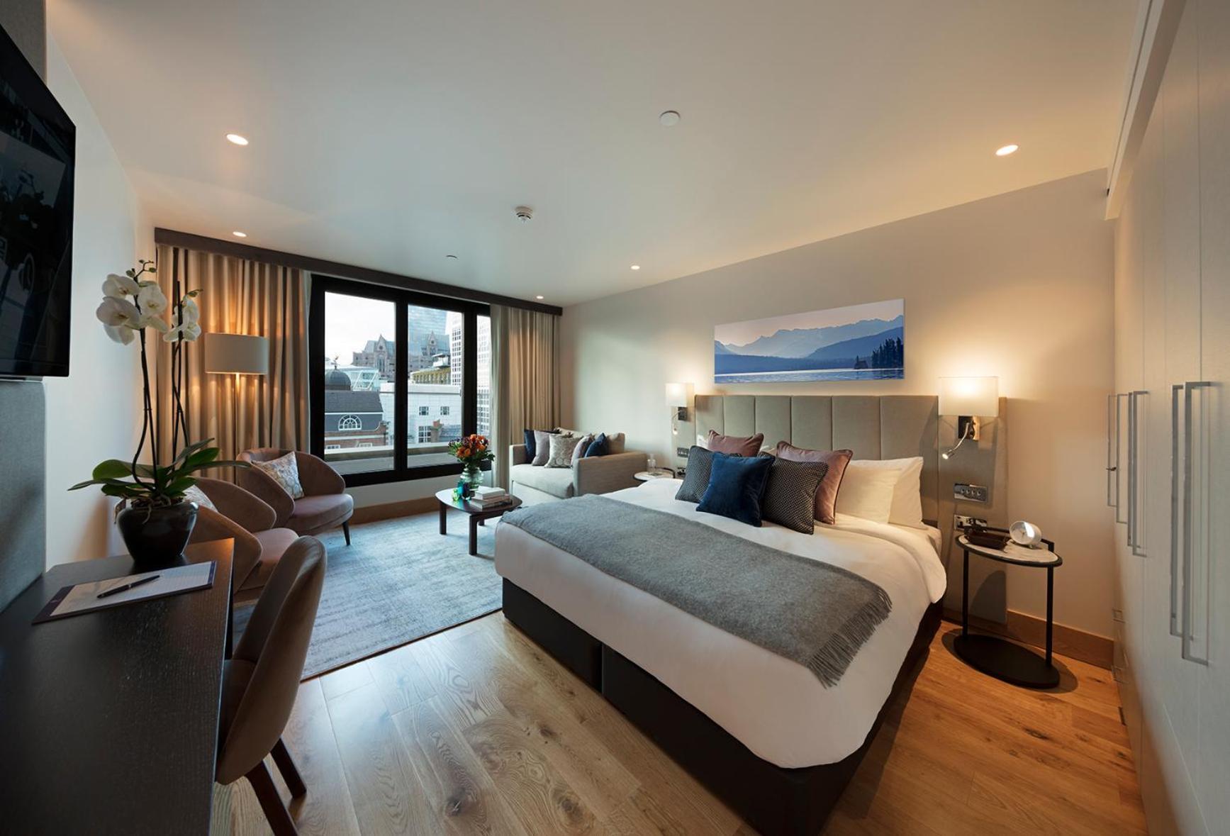 Tower Suites By Blue Orchid لندن المظهر الخارجي الصورة The photo shows a modern hotel room. In the center, there is a large bed with stylish bedding and pillows in various shades. To one side, there are two comfortable chairs and a small table, creating a cozy seating area. A large window offers a view o